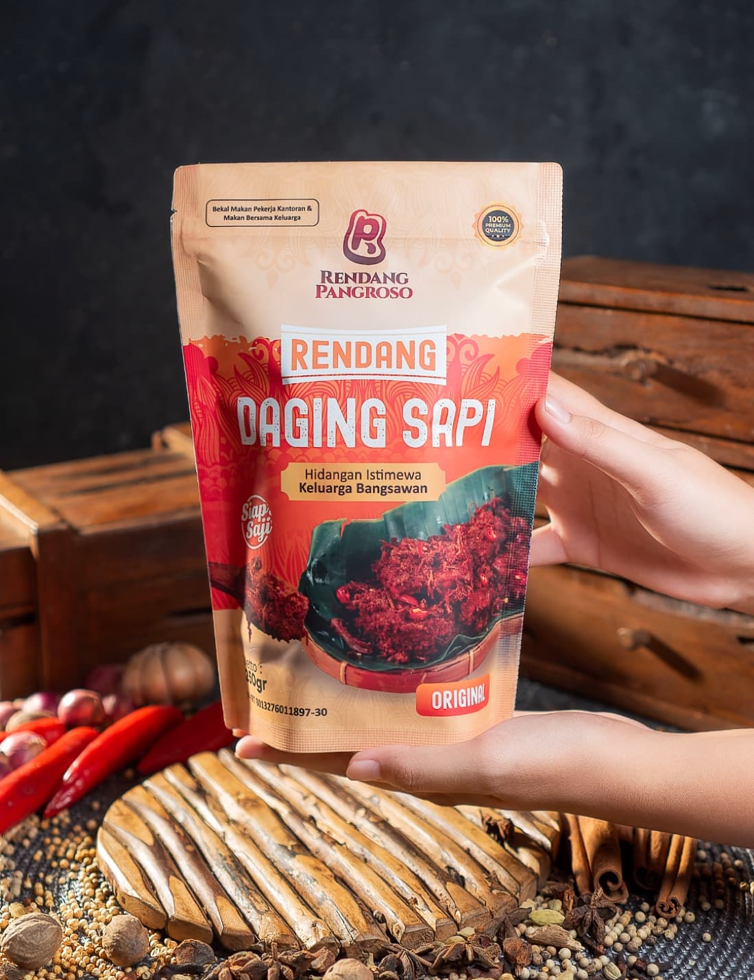 Kemasan Rendang Pangroso dipegang di atas latar kayu dan rempah